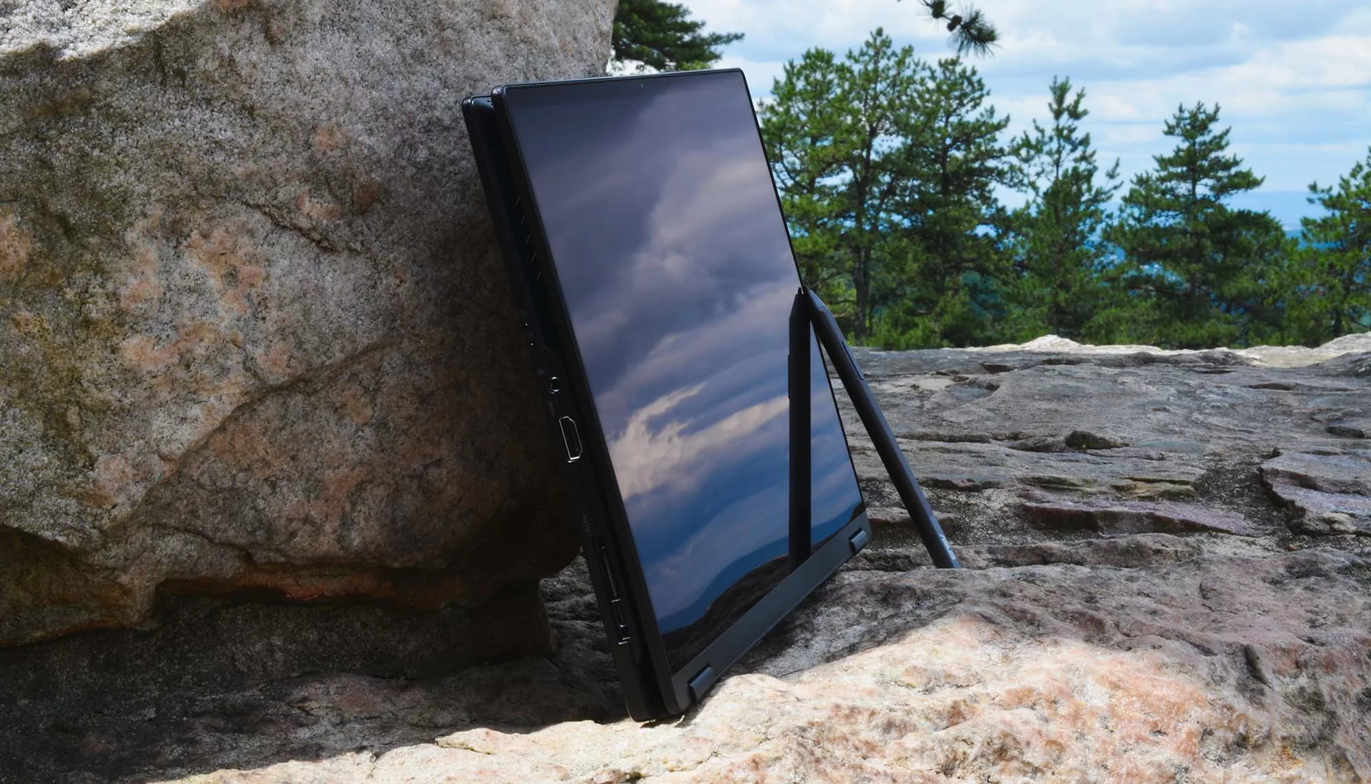 X13 in tablet mode with a stylus, leaning against a boulder with trees in the background.