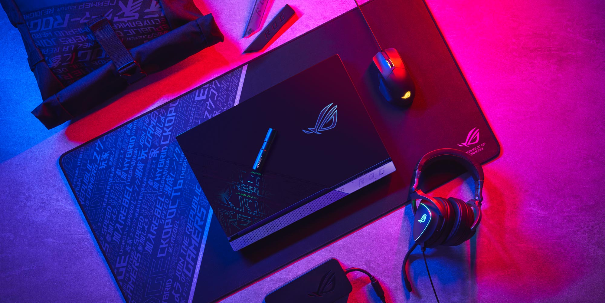 An image of the ROG Strix SCAR 17 SE sitting on a desk mat, with the lid closed, surrounded by a mouse, backpack, and charger.