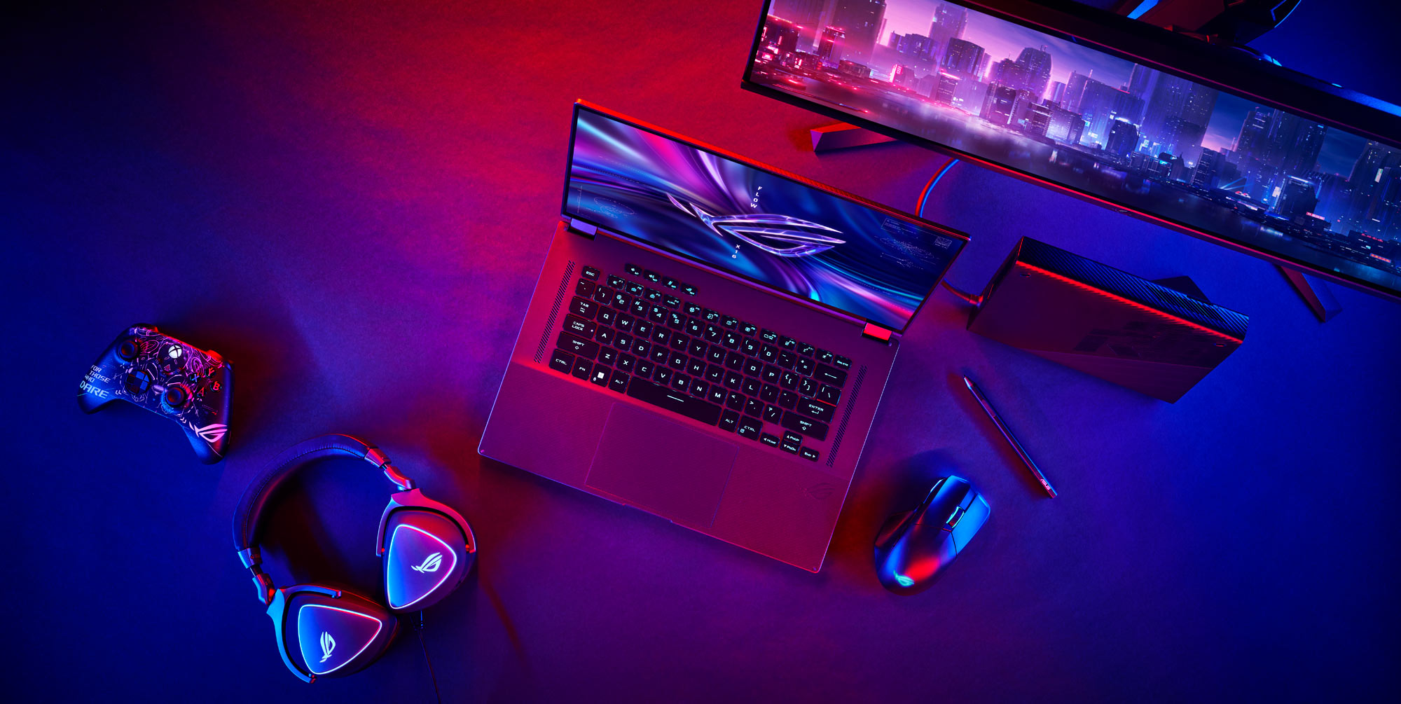 A birds-eye view of the ROG Flow X16 laptop, next to a mouse, controller, headset, and monitor.