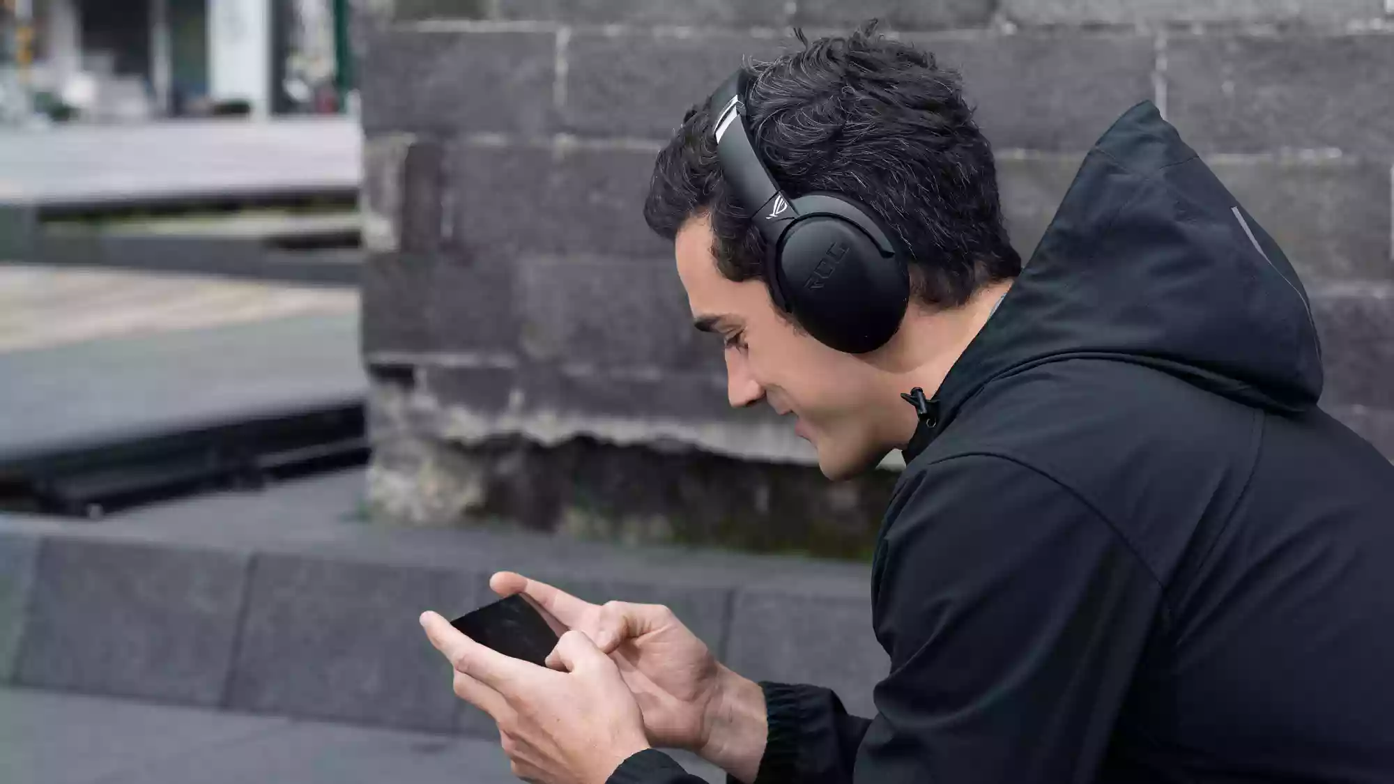 A man wearing ROG Strix Go BT headphones playing games on an ROG Phone outside on the street.
