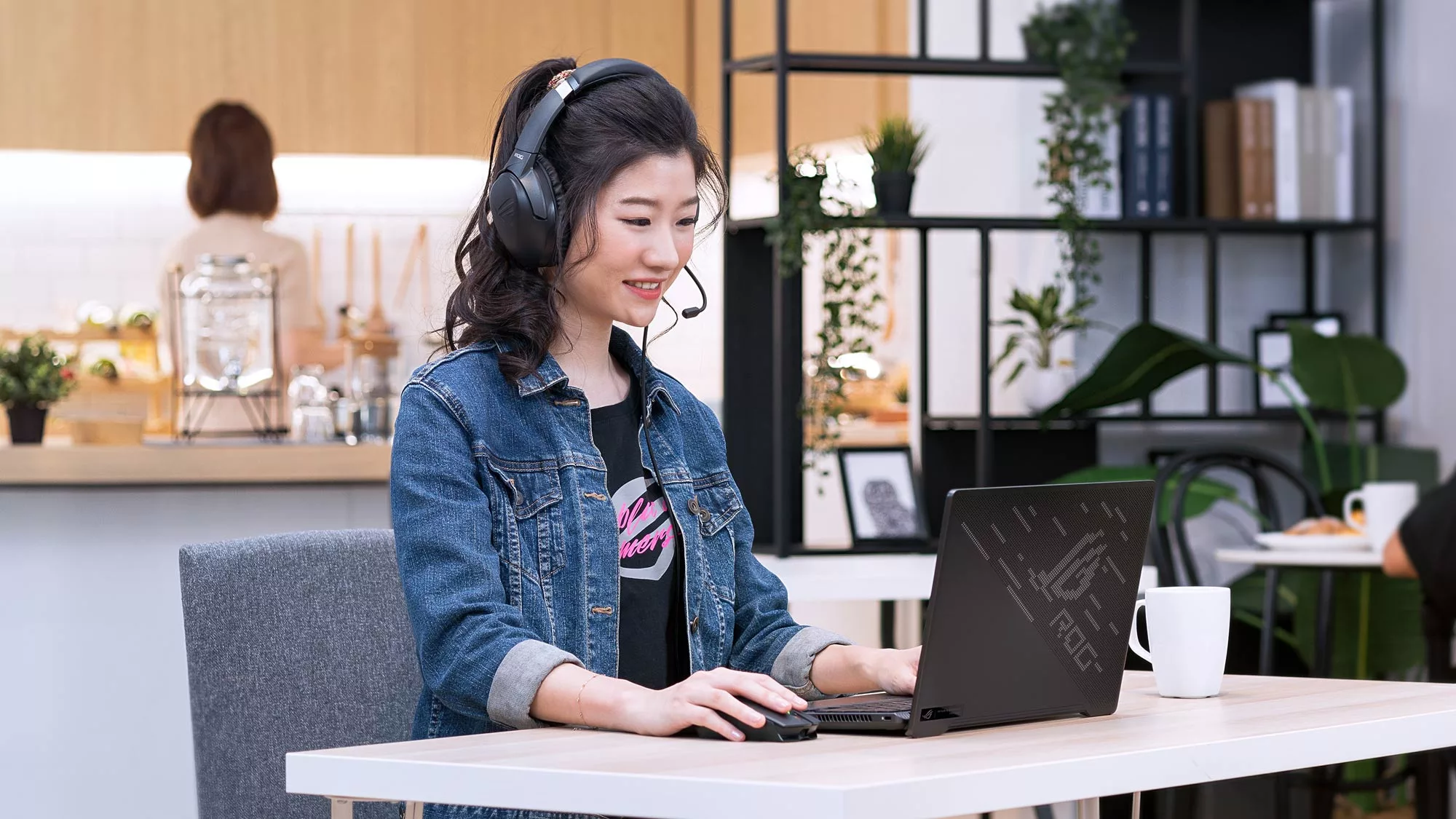 A smiling woman sitting at a table with an ROG laptop wearing the ROG Strix Go headset.