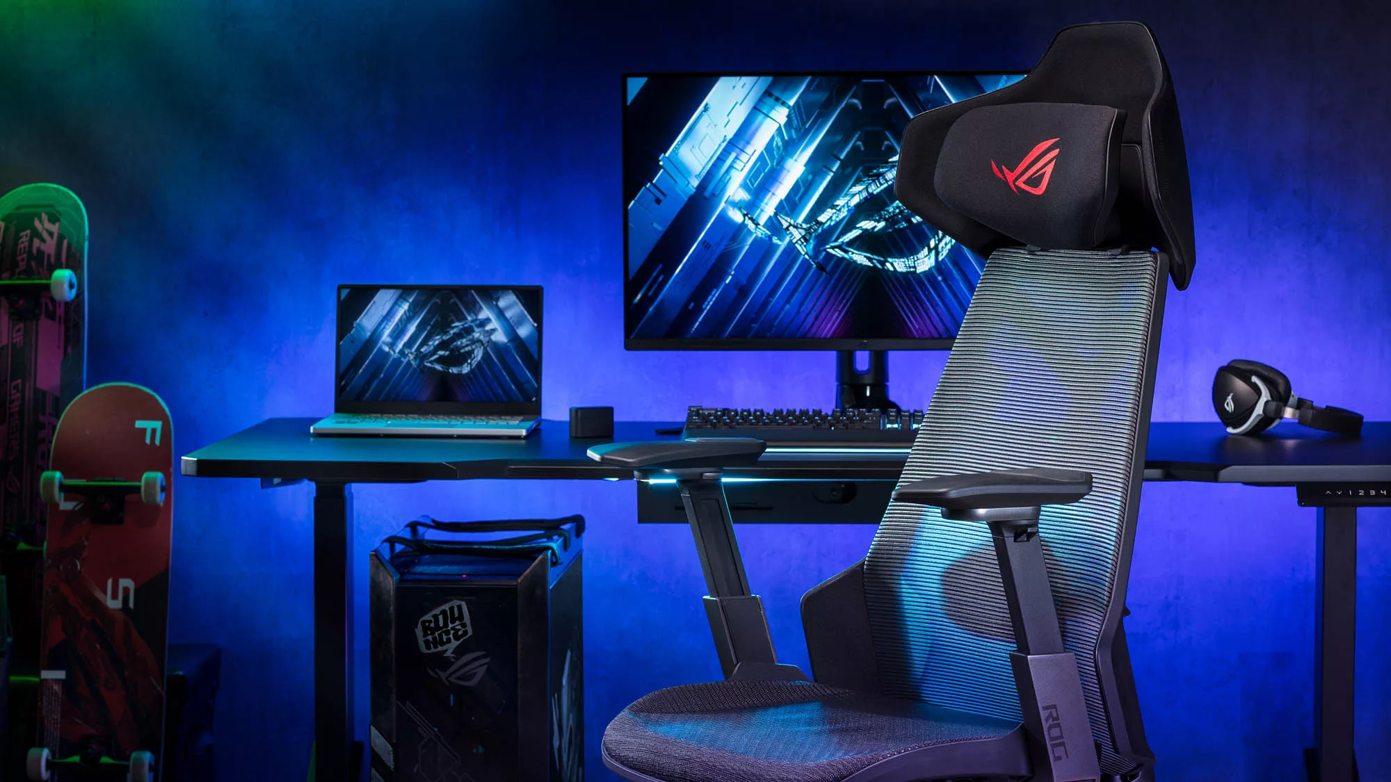 An ROG laptop and desktop setup alongside a pair of ROG headphones, and ROG chair, and skateboards against a purple wall.